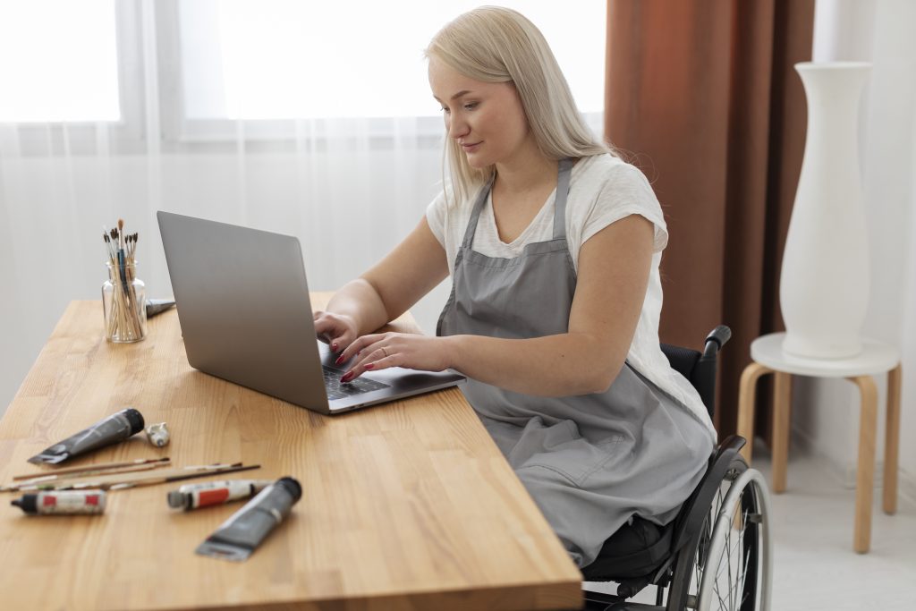 Female wheelchair user at a desk. She has long blonde hair, is wearing a white top and grey apron. She looking at, and typing on a laptop scree. Behind her is a window.