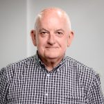 An older man with a black and white checkered shit on sits at the centre of the impage agains a light plain bakcgound.