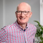 A man is wearing a red,blue and white checkered shirt as he leans int the image. He is bald with no facial hair and is wearing black rectangular glasses.