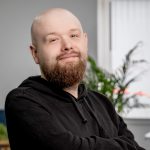 A man is sat in an office. He is bald with a thick ginger and white beard. He has soft eyes that are blue and is wearing a black hoodie.