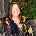 a woman i sat agains a tree trunk. She is laughing with a wide smile. She has straight mid-legnth hair and black and yellow blouse on.
