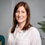 Florence is a white woman with shoulder legnth dark hair. She has blue eyes and a wide smile. She is in an office setting and wearing a louse fit white blouse.