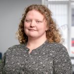 A young woman with light, shoulder legnth very curly hair. She is wearing a dark blue and earthy green floral shirt and is wearing small stud earrings.