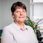 A woman with short brown hair is sat in an office with a plant behind her. She has a grey shirt on and a neutral expression.