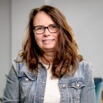 A woman wearing a denim jacket and whit etop sits in a plain room. She has long brown hair and is wearing dark brown glasses.