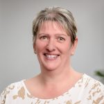 A white woman with short blonde hair. She is wearing pearl earrings and a white and beige oatterend top.