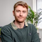 A white, young man with ginger hair and a short beard is sat within an office. There is one blurred plant behind his right shoulder. He has blue eyes and is wearing a forest green jumper over a light green shirt. He has a small gold hoop earing at thetop of his right ear.