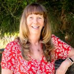 A middle-aged woman with a bright red and white floral t-shirt on stands outdoors, in front of a tree. She has long hair that is brown at the root and blonde at the ends. She has a very friendly and welcoming smile amd experssion.