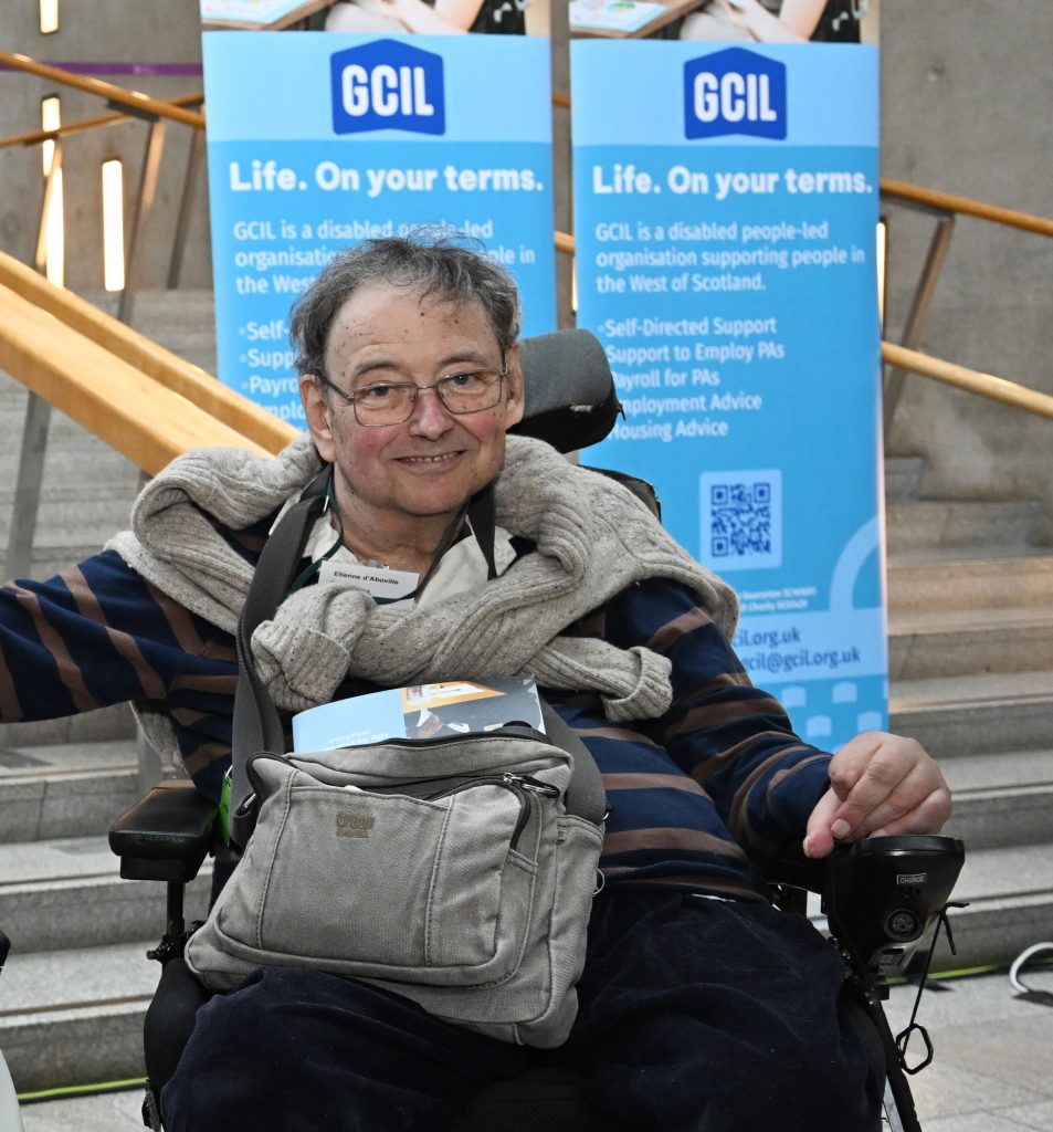 Man sitting down. He has a beige jumper tied around his neck and a satchel on his lap. He is wearing glasses and smiling at the camera. He is sitting in a wheelchair. Behind him is a set of stairs, and two pull up banners showing some text and the logo for GCIL.