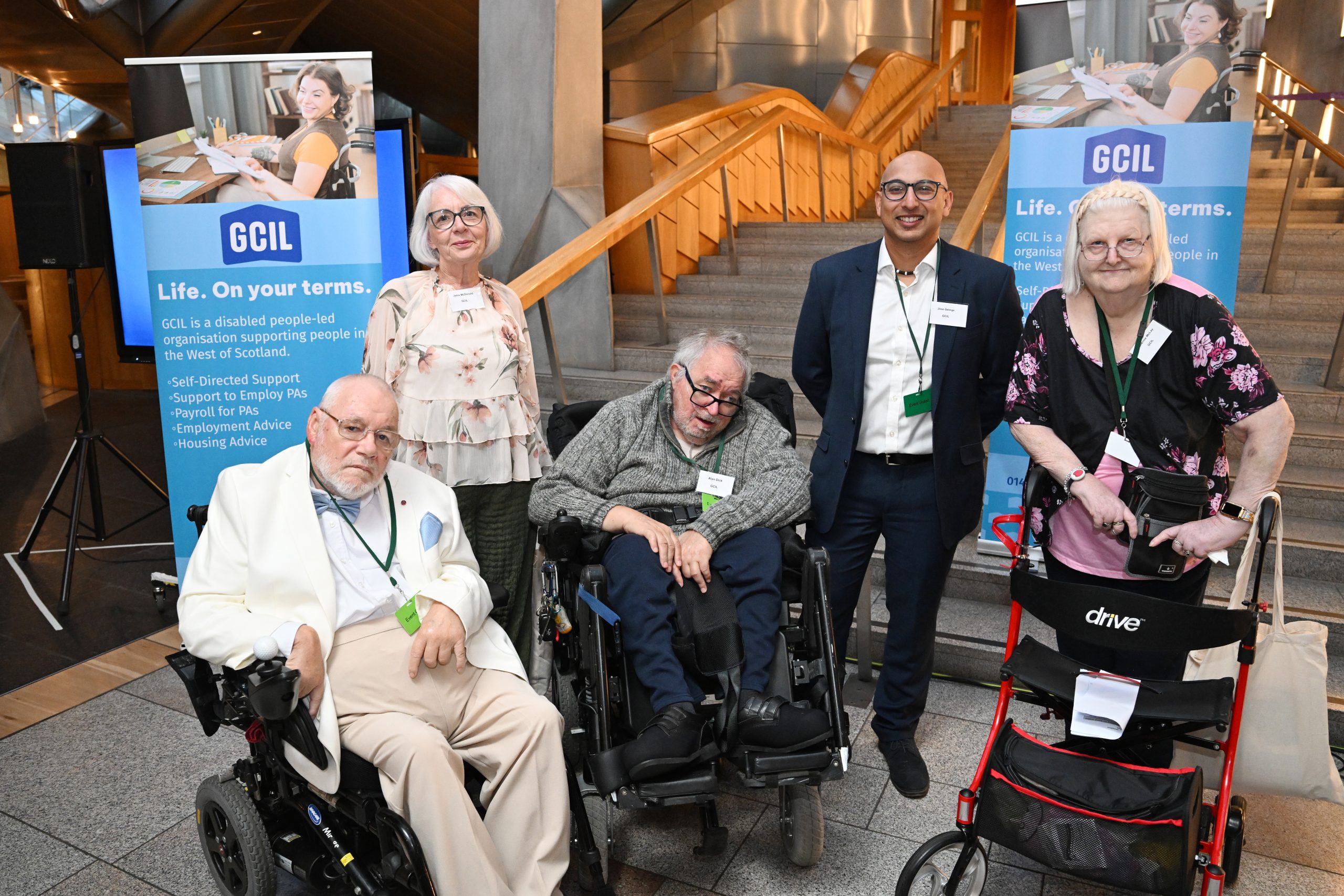 A group photo of 5 people. From left to right; a man sitting in his wheelchair. He is wearing a cream jacket and beige trousers, white shirt and blue bow tie. He has glasses and balding head. Then a lady standing behind. She is wearing a floaty flowery blouse, has grey bobbed hair, glasses. Then in a man sitting in his wheelchair, wearing a grey jumper, dark blus trousers and he is wearing glasses and has grey hair. Next along in a man standing wearing a dark blue suit and white shirt. He is wearing glasses and is bald. Finally a lady stading leaning on a walking frame. She is wearing a black and pink flowery top, has white shoulder length hair and is wearing glassses. The group are in a building. Behind them is a set of stime stairs, wooden handrails and a pull up banner showing the logo for GCIL.
