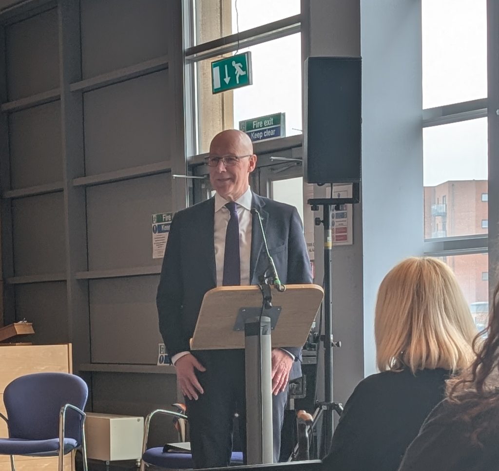 Image of the Scottish First Minister John Swinney. He is standing at a lectern and microphone. He is wearing a suit and tie. Behind him are windows grey wall and blue chairs.