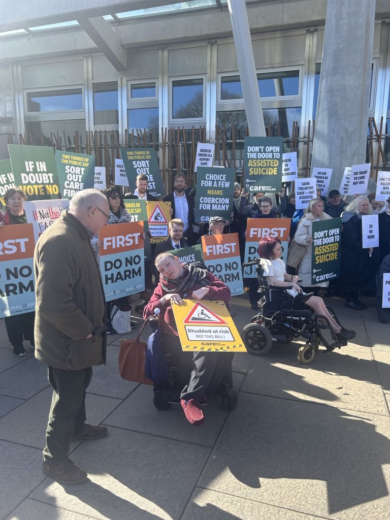 The image is of many people standing outside a building. They are holding placards. Placards show slogans such as First do no harm, and If in doubt vote no. Some individuals are sitting in wheelchairs, some are standing.
