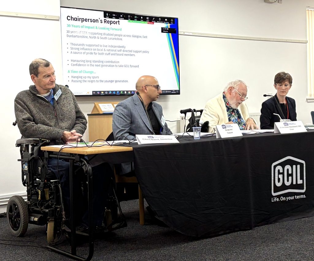 A panel of four individuals sits behind a long table covered with a black cloth displaying the GCIL logo and the slogan “Life. On your terms.” One panelist uses a powerchair. Each person has a microphone and nameplate in front of them, suggesting a formal meeting or annual gathering. Behind the panel, a large projected slide titled “Chairperson’s Report” summarizes GCIL’s 30 years of supporting disabled people across Glasgow and surrounding regions, highlighting achievements such as independent‑living support, policy influence, and staff and board pride. The slide also notes a leadership transition, with references to “hanging up my spurs” and passing responsibilities to a younger generation. The room appears to be a conference or organizational meeting space, with the panel focused on presenting the report to attendees.