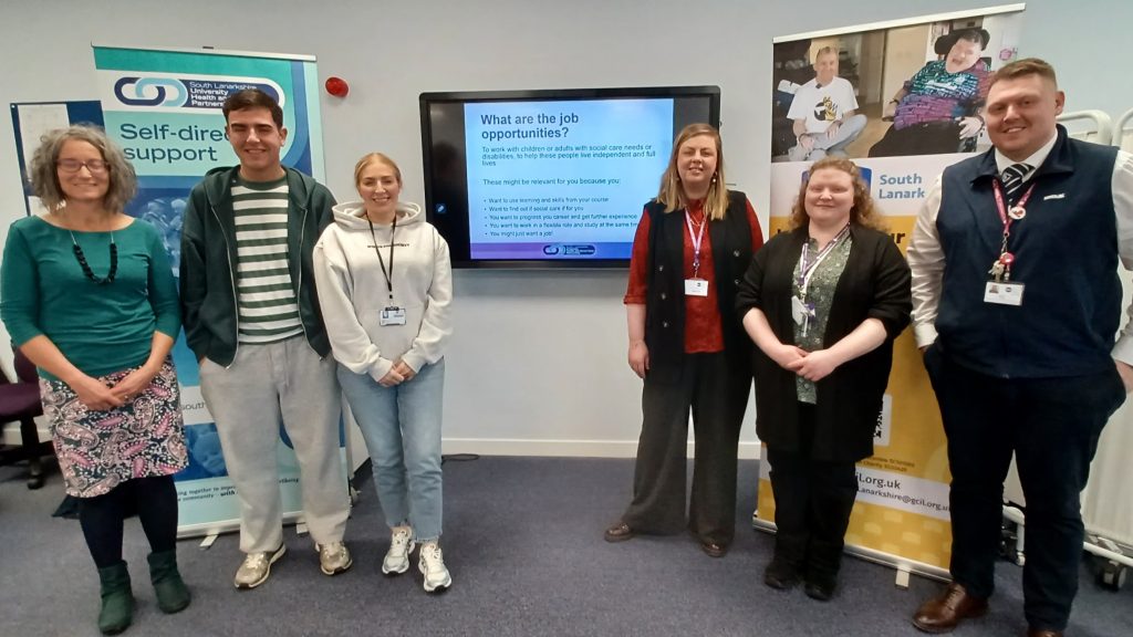 Image of 6 people standing in a line. Between them in the middle is a tv screen on the wall which a display reading What are the job opportunities (and other unreadable text). Behind the people on each side are also 2 pop-up banners advertising Self-directed support and South Lanarkshire council. The people are all smiling. There are 4 women, dressed casually and 2 men. They are a range of ages and heights.