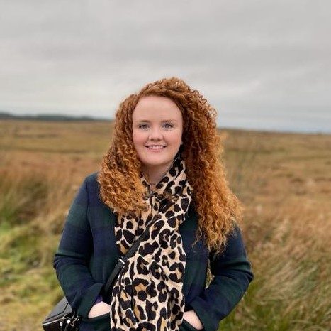 Photo of red head stood in a field with her hands in her pockets and smiling.