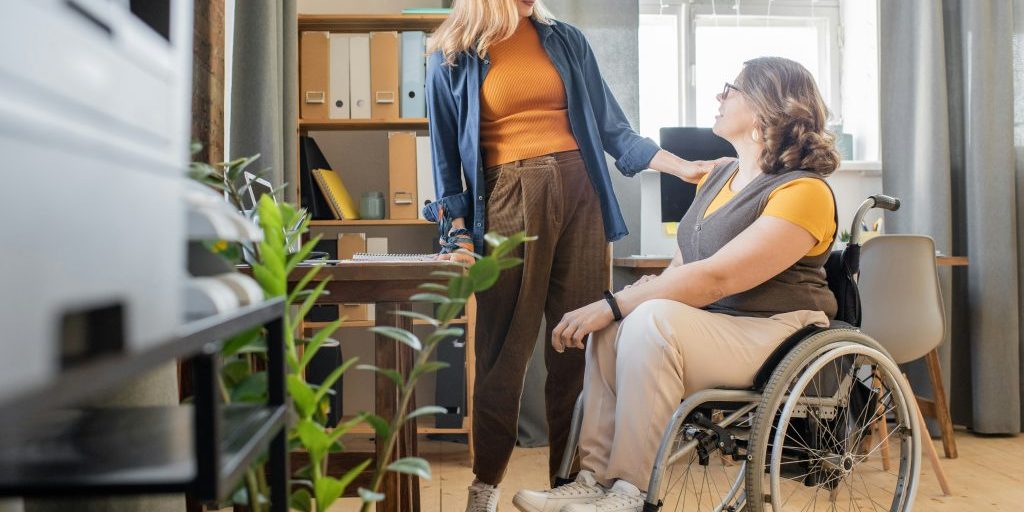 Woman standing beside another woman in a wheelchair looking at each other.