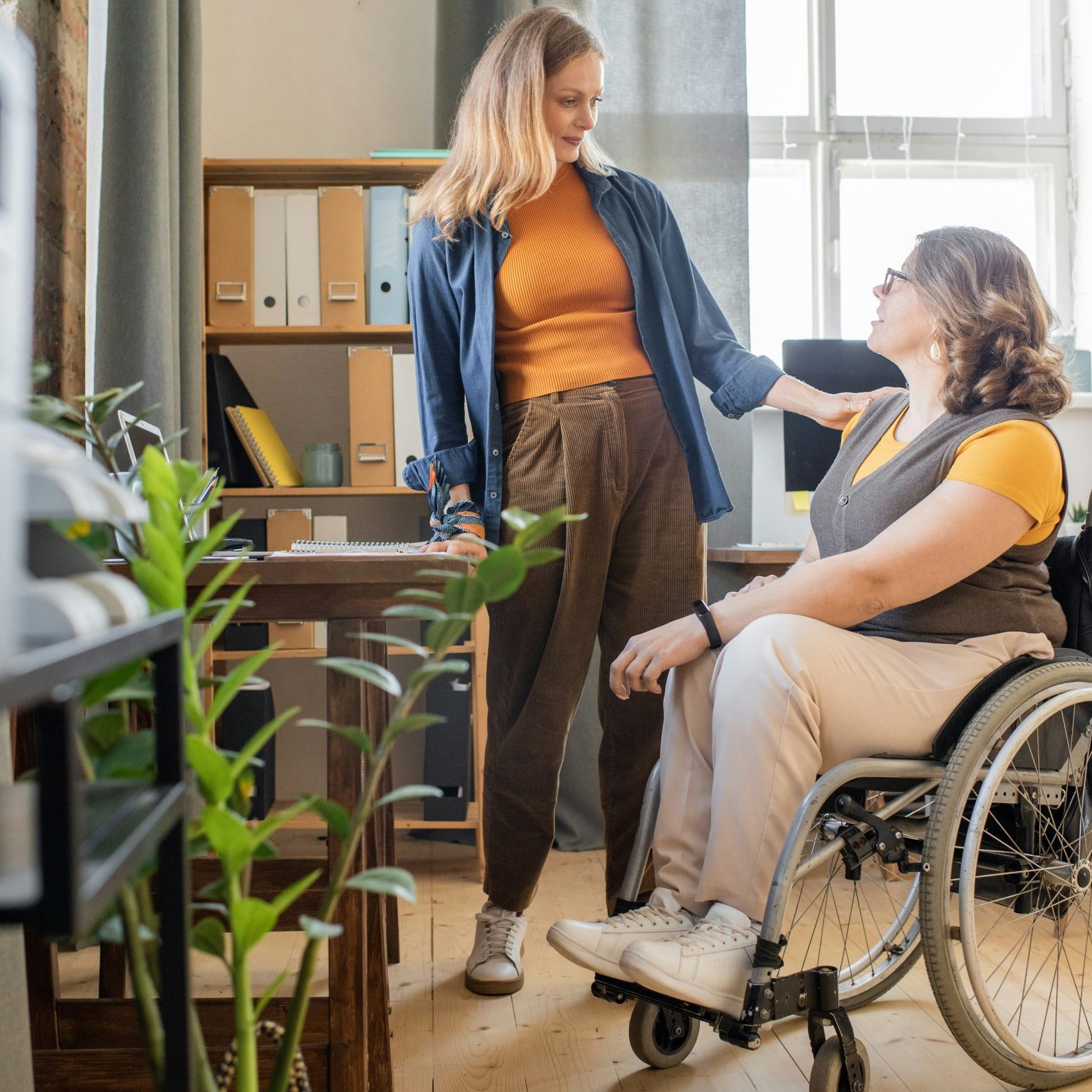 Woman standing beside another woman in a wheelchair looking at each other.