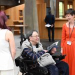 A man in a wheelchair smiles and holds a booklet while talking with two women indoors. One wears a white outfit, the other an orange dress. The mood is friendly.